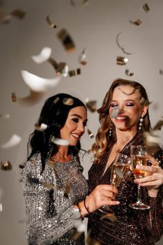 This may contain: two beautiful women standing next to each other holding wine glasses with confetti flying around them