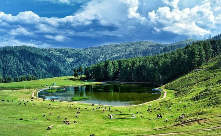 This may contain: a lake surrounded by green grass and mountains in the distance with people walking around it