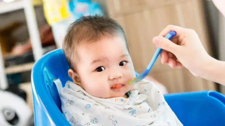 A baby sitting in a blue high chair while being fed with a spoon by an adult hand.