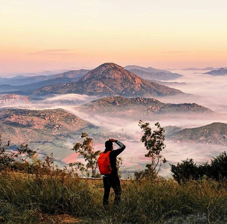This may contain: a man standing on top of a lush green hillside next to a mountain covered in clouds