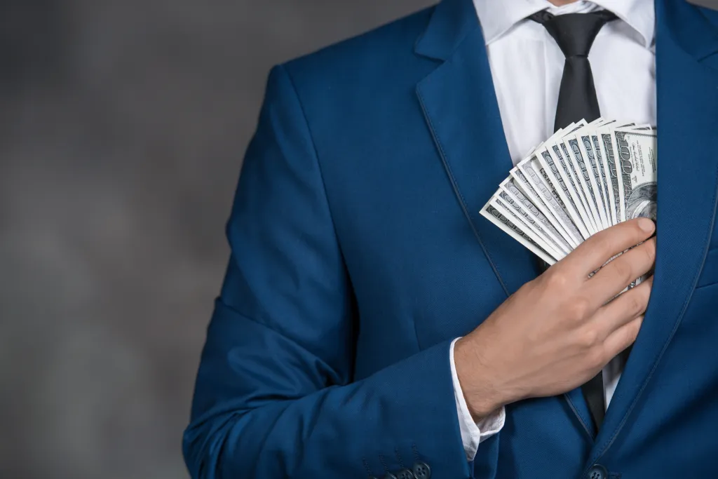 Man in a blue suit and white shirt putting a fan of hundred-dollar bills into his inner jacket pocket.
