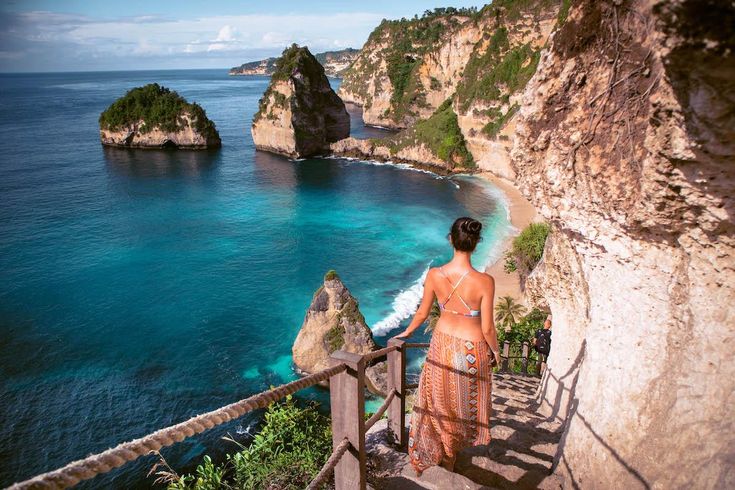 This may contain: a woman walking up some stairs towards the ocean and cliffs with blue water in the background