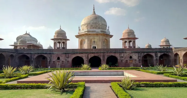 A historic Mughal-style monument with multiple domed pavilions rising above an arched colonnade, set behind a symmetrical garden with trimmed hedges under a clear sky.