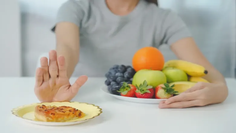 A person in a gray shirt pushes away a plate of pizza, opting for a plate of fresh fruits like apples, bananas, grapes, and strawberries, signaling health-conscious choice.