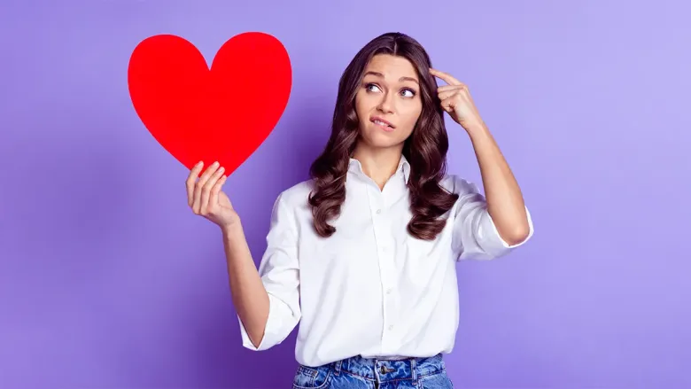 A thoughtful or confused woman holding a large red heart cutout. 