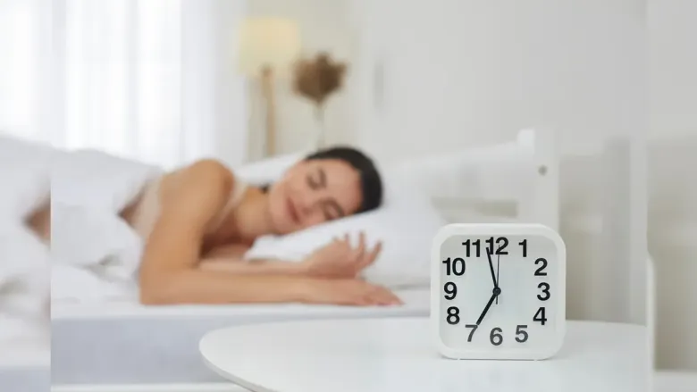 A woman sleeping in a bed with a white alarm clock in the foreground showing 7:00. 