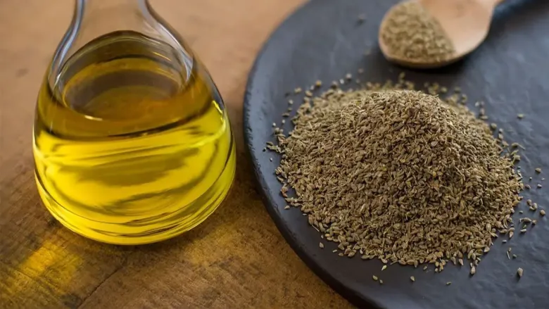 A small glass container filled with yellow oil sits on a wooden surface beside a dark slate plate holding dried ajwain seeds and a wooden spoon.