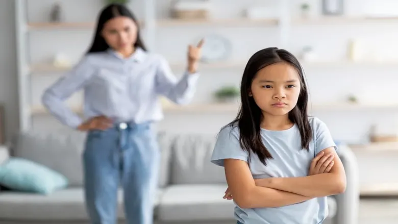 a girl is standing with folded hands in a serious mood with mom talking in background