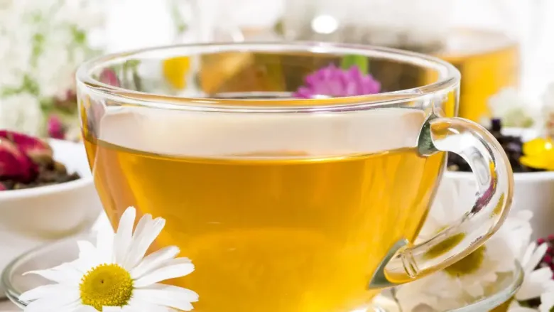 A clear glass cup filled with light golden herbal tea, placed on a glass saucer with white daisy flowers around it. The background shows blurred flowers and tea ingredients, creating a fresh and soothing atmosphere.