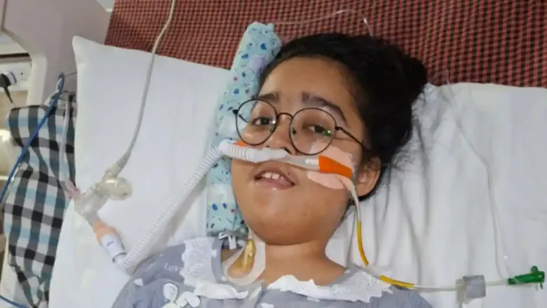 A close-up portrait of Kanishka Bisht lying in a hospital bed. She is wearing round glasses and has a breathing tube (BiPAP mask) secured to her face to assist with her respiratory condition.