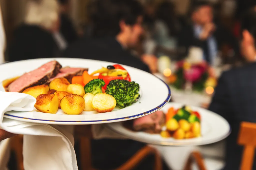A waiter carries two plates of beef, roasted potatoes, broccoli, and carrots at an event.