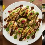 Top with tamarind sweet chutney, green chutney, chaat masala, and red chili powder, then garnish with sev, pomegranate seeds, and fresh coriander before serving.