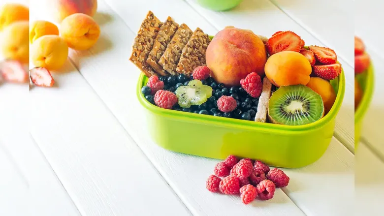 A green bento box filled with fresh fruit and seed crackers on a white wooden table.