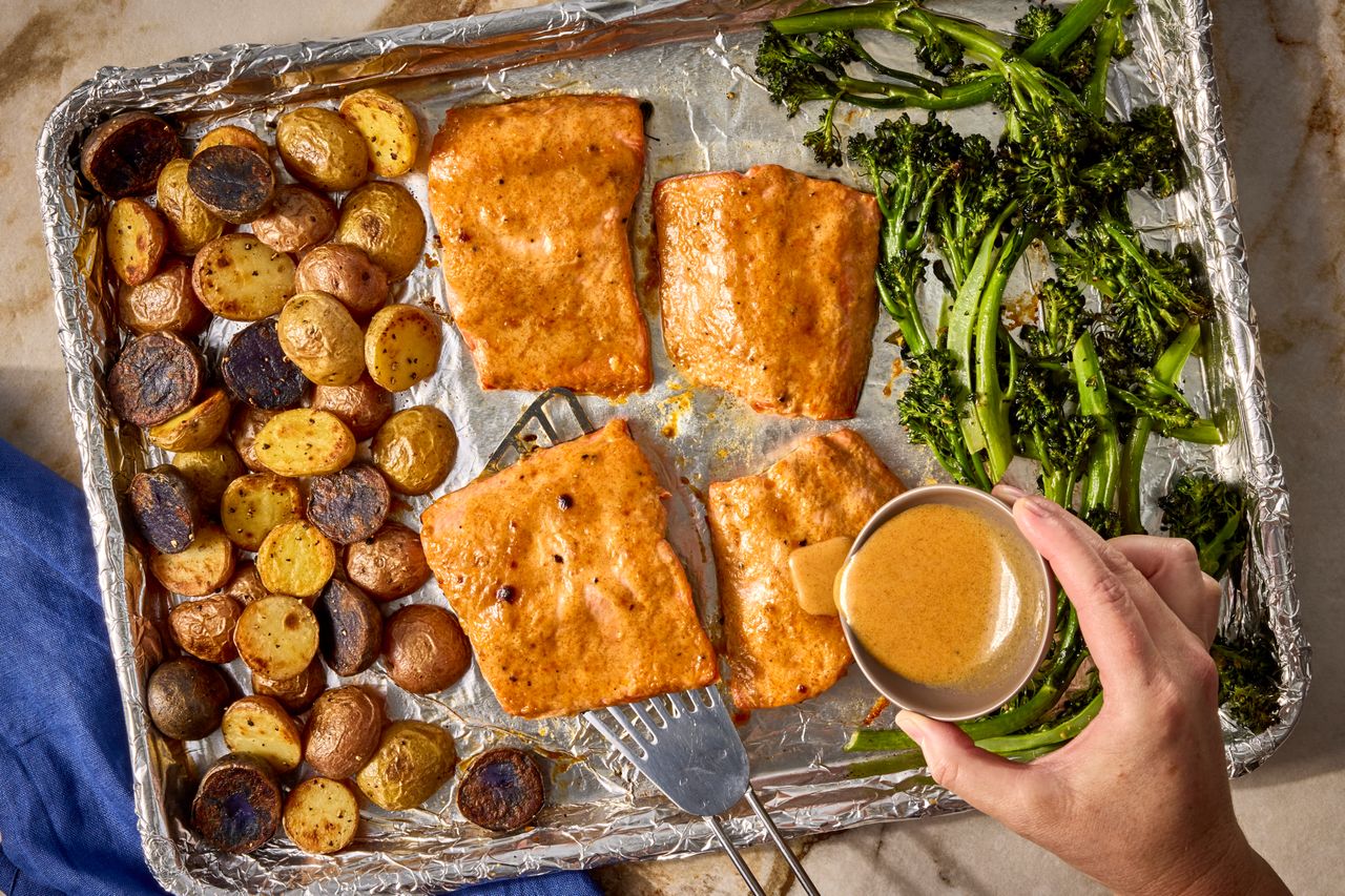 Baked salmon filets on a sheet pan with roasted potatoes and broccolini a hand drizzling sauce over the salmon Credit: Photographer: Jen Causey, Food Stylist: Emily Nabors Hall, Prop Stylist: Hannah Greenwood.