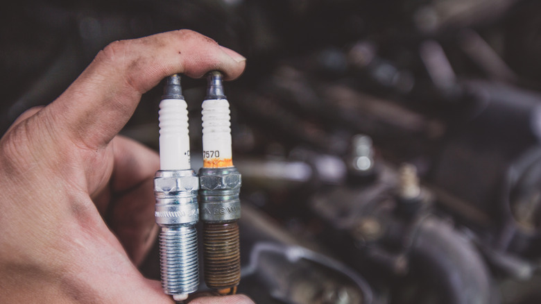 A man holding a new and old spark plug side-by-side, out of focus engine in the background