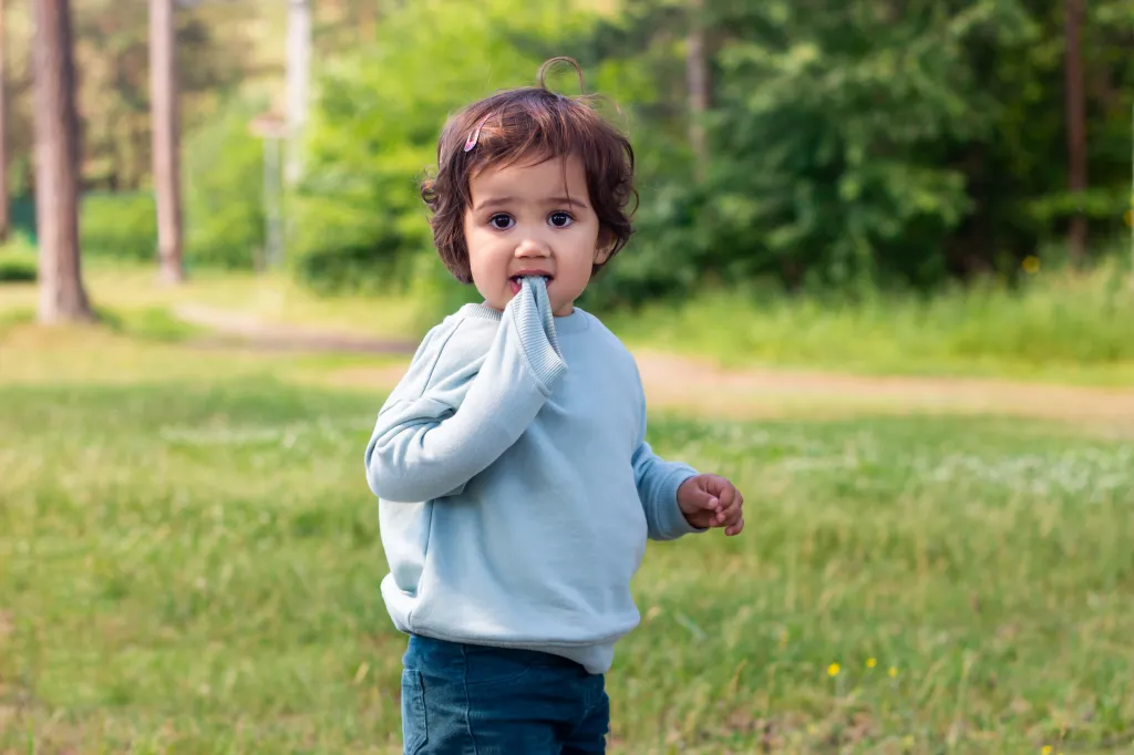 A young child stands in a grassy park, chewing on the sleeve of a light blue sweatshirt.