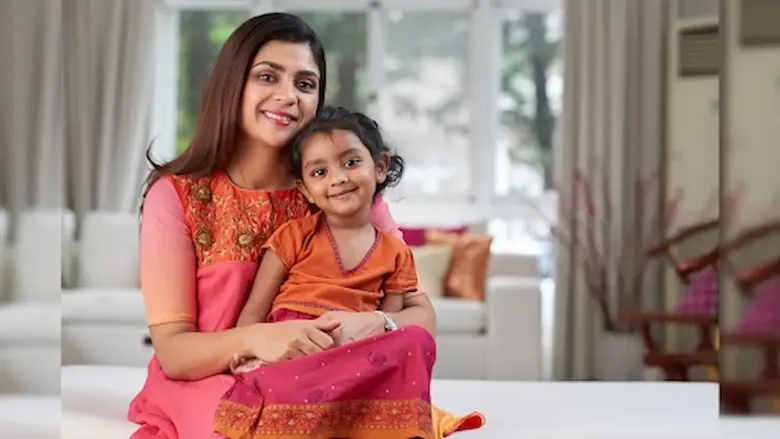 A smiling Indian woman and her young daughter, both in colorful traditional attire, sit together in a brightly lit room.