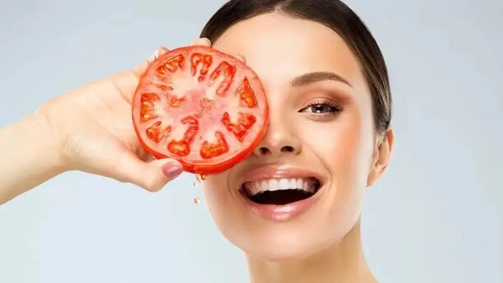 Smiling woman holding a slice of tomato over one eye, symbolizing natural skincare and glow