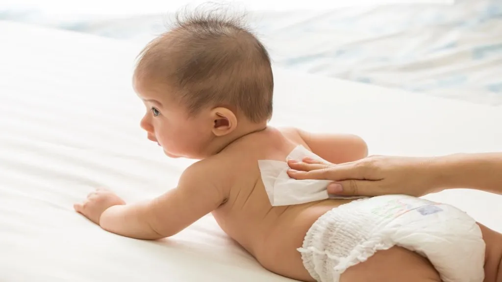 A baby lying on their stomach wearing a diaper, while an adult hand gently wipes the baby's back with a wet wipe.