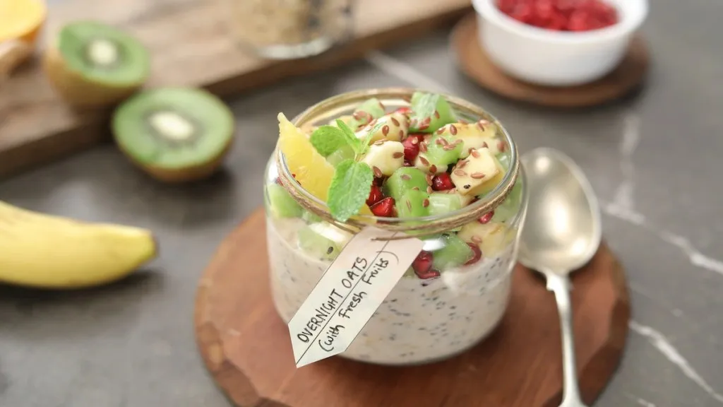Jar of overnight oats topped with kiwi, banana, pomegranate, seeds, and mint, served on a wooden board.