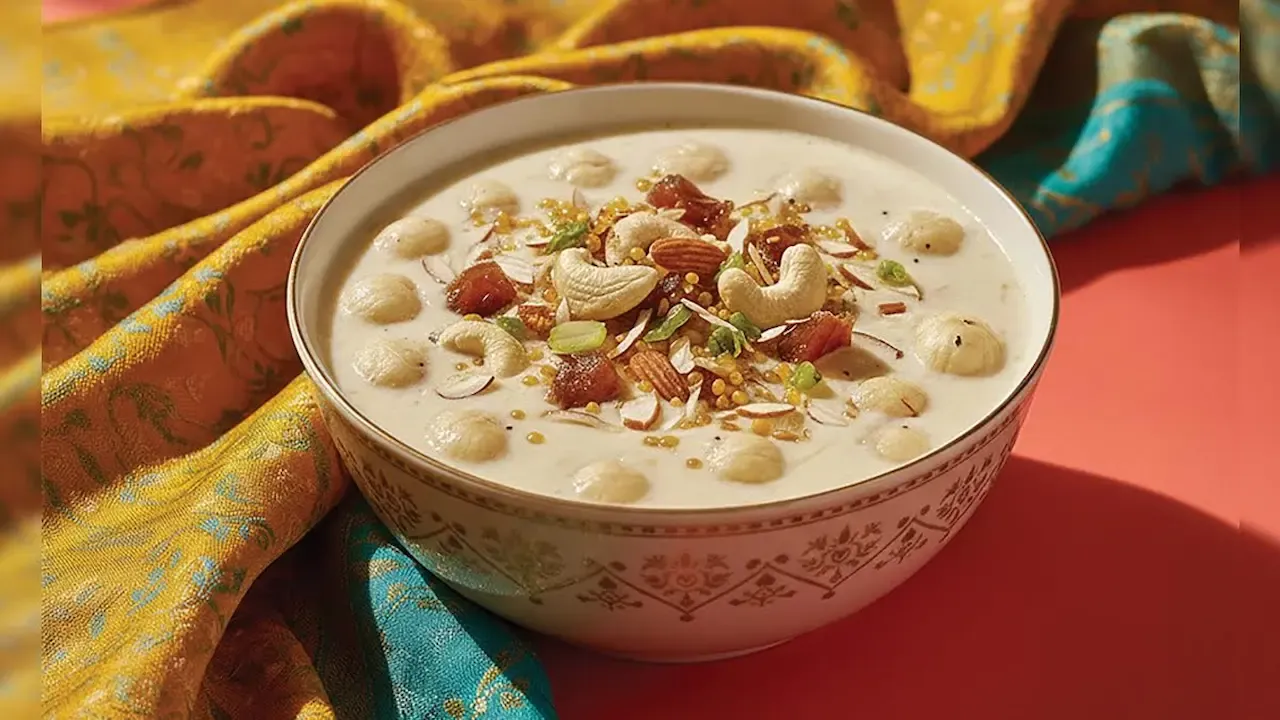 Bowl of creamy Indian dessert garnished with nuts, dried fruits, and seeds, placed on a colorful festive cloth.