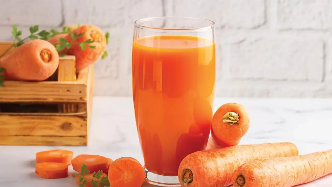 Glass of fresh carrot juice with whole and sliced ​​carrots placed around on a kitchen surface.