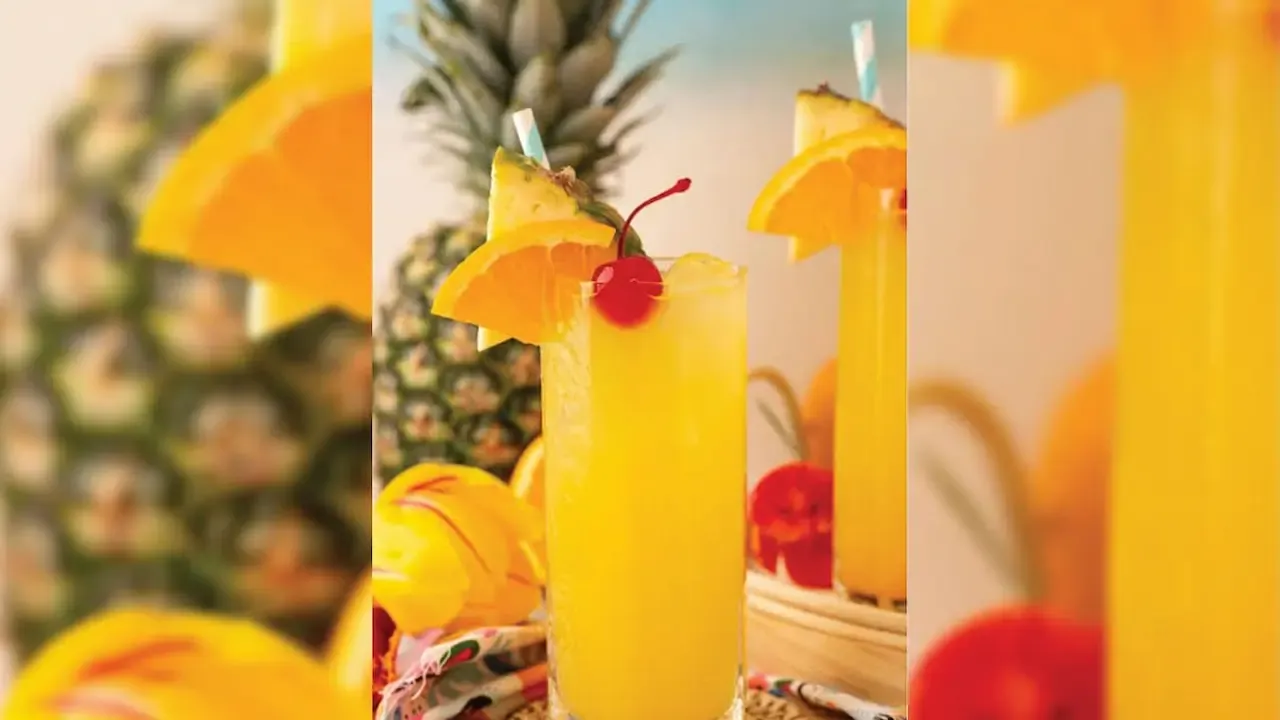 Bright yellow tropical drink in a tall glass, garnished with pineapple slice, orange wedge, cherry, and straw.