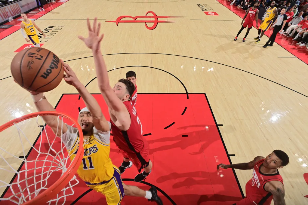 A Lakers player in a yellow jersey with number 11 is mid-air about to dunk a basketball, while a Rockets player in a red jersey attempts to block.
