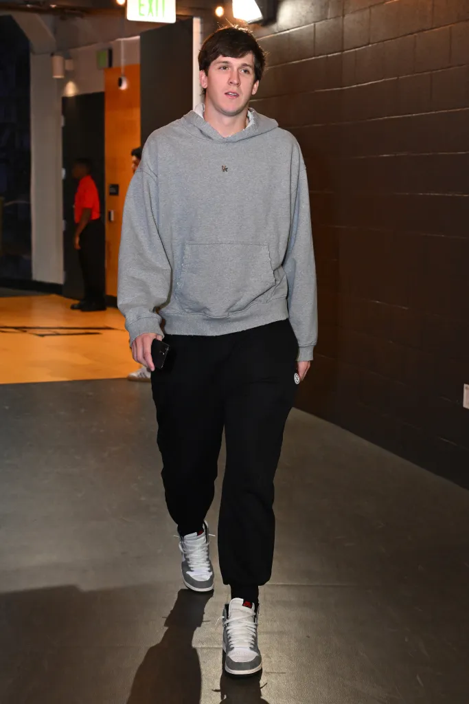 Austin Reaves walking in a hallway, dressed in a grey hoodie, black pants, and white and grey sneakers, holding a phone in his left hand.