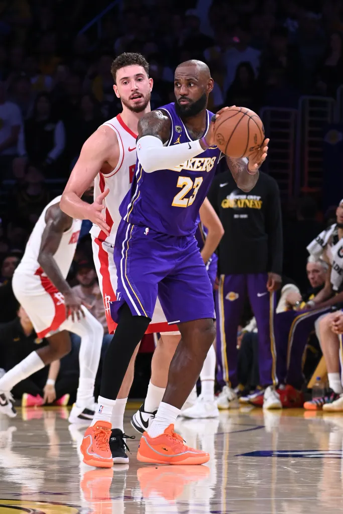LeBron James, wearing a purple and yellow Lakers uniform with number 23, holds a basketball, guarded by a Houston Rockets player in a white and red uniform.