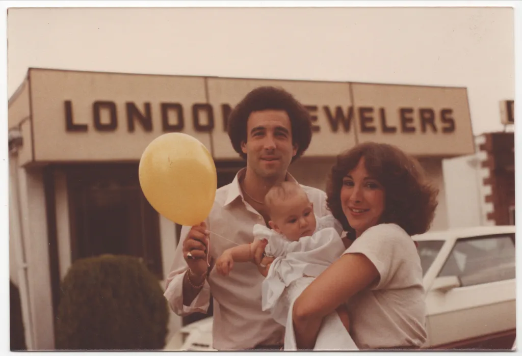 A man holds a yellow balloon and a baby as he poses next to a woman in front of a "London Jewelers" sign.