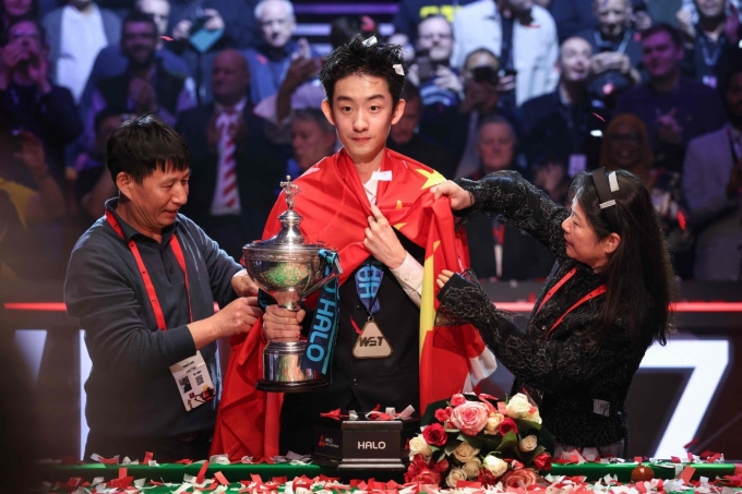 Wu Yize (C) holds the trophy as his parents wrap a Chinese flag around his shoulders after his victory over Shaun Murphy in the World Snooker Championship final in Sheffield, England on May 4, 2026. Photo by AFP
