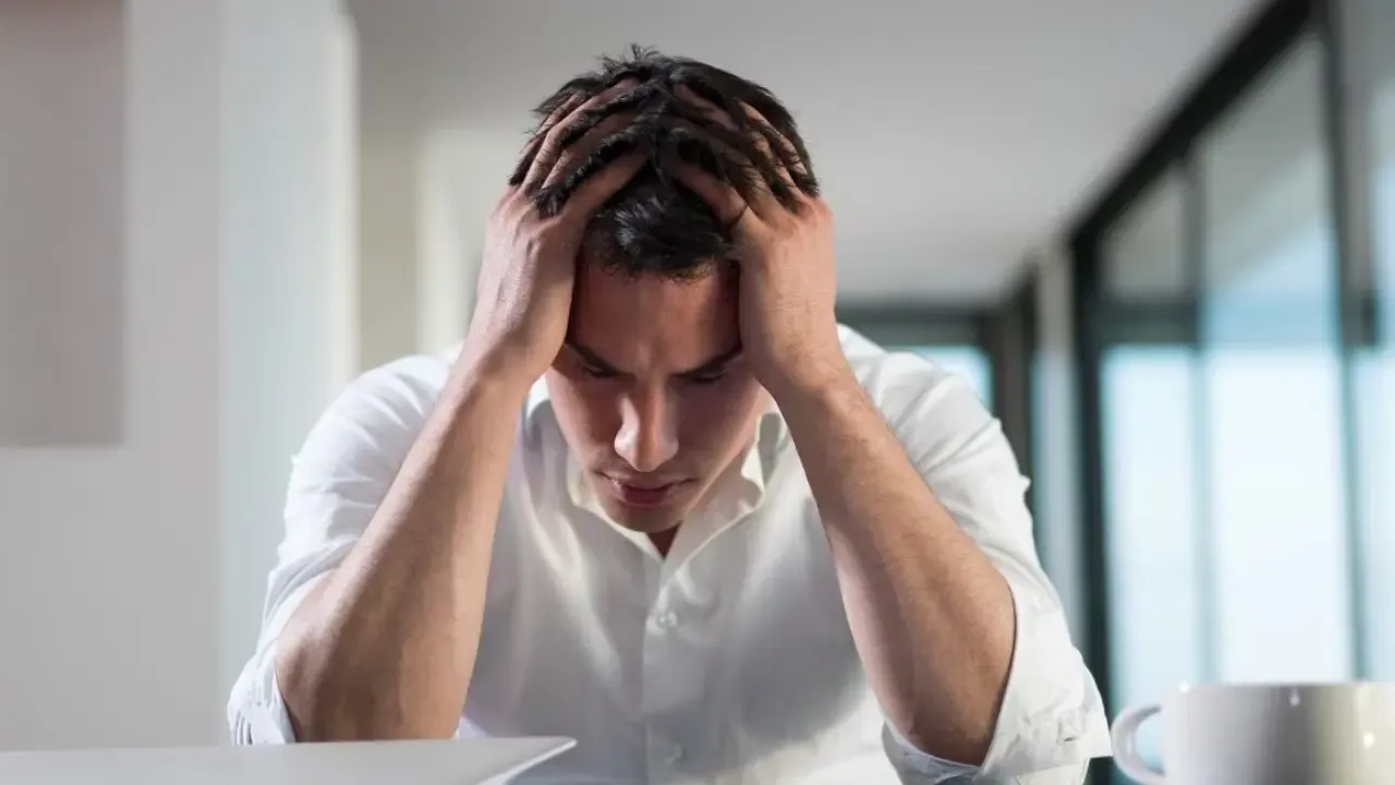 A man in a white shirt sits with his head in his hands, looking down in stress or frustration, with a laptop and a cup placed on the table in front of him.