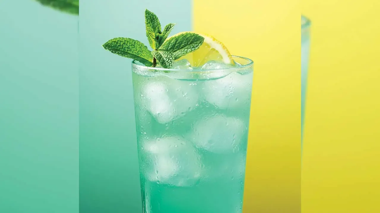 A tall glass filled with an icy teal mint mojito, garnished with a lemon slice and a fresh mint sprig on a green and yellow background.