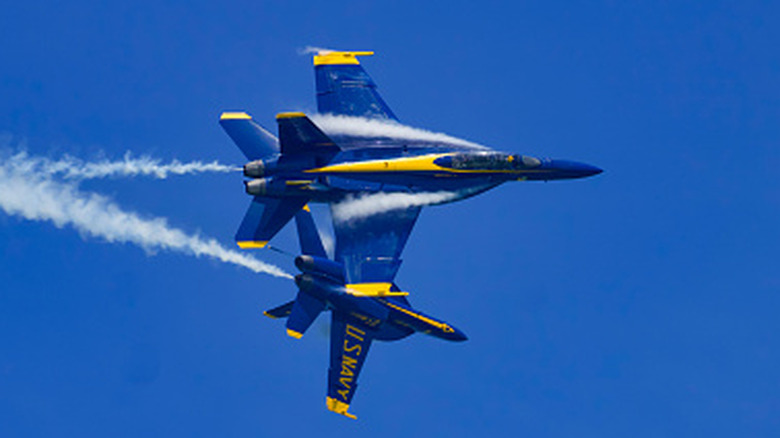 Two U.S. Navy Blue Angels jets fly in tight formation against a clear sky, leaving white smoke trails as they perform an aerobatic maneuver.
