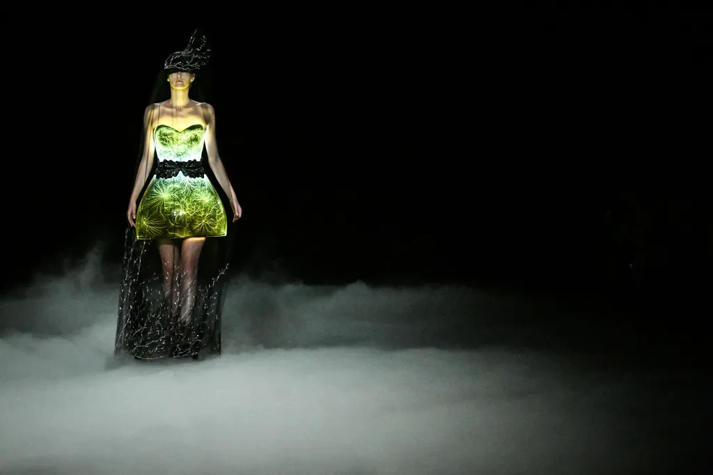A model presents a strapless green and yellow dress with a lightning-like pattern and a dark, ornate headpiece, standing in a cloud of fog.