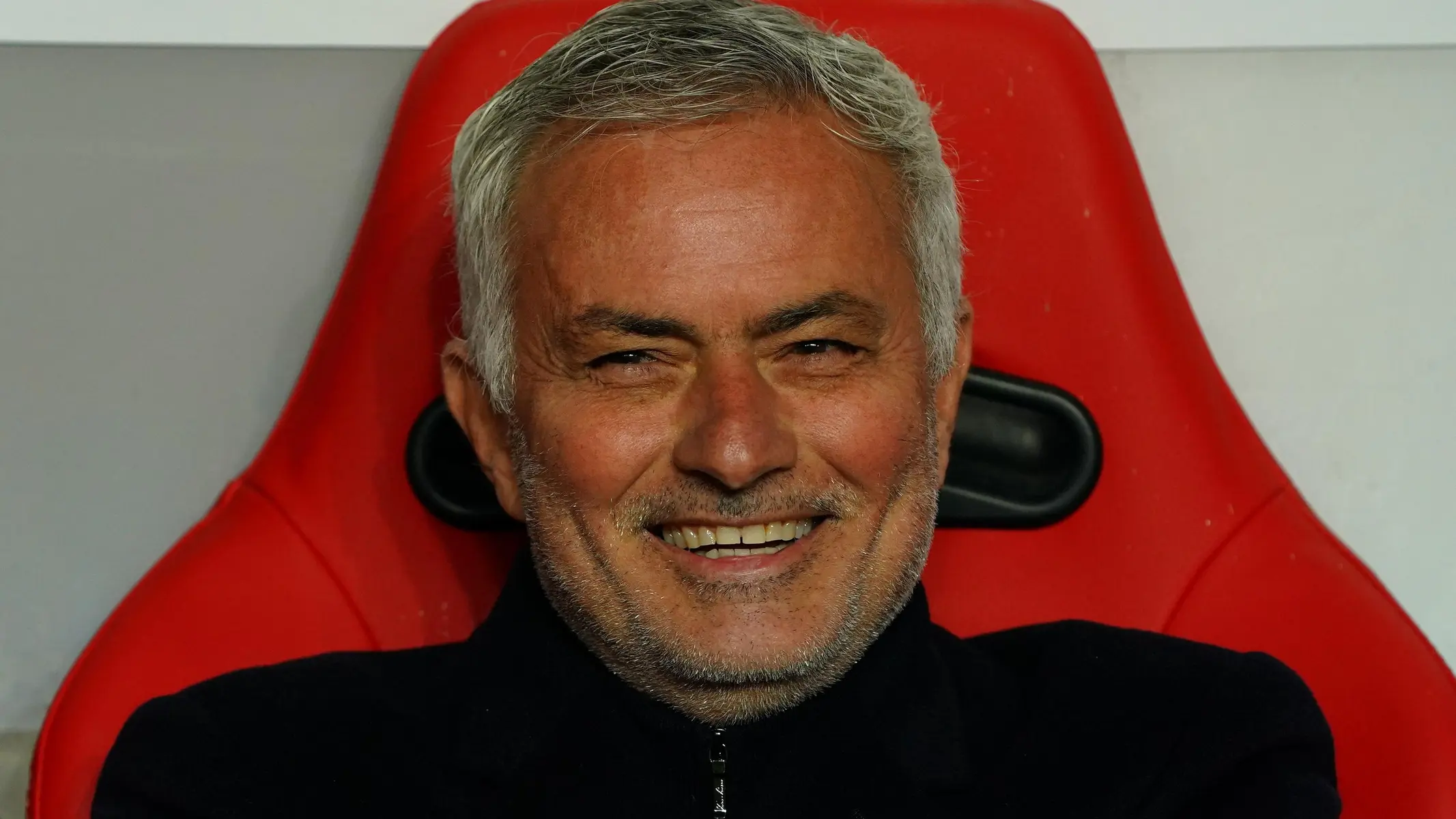 Jose Mourinho smiling while seated on a bench in a sports setting.