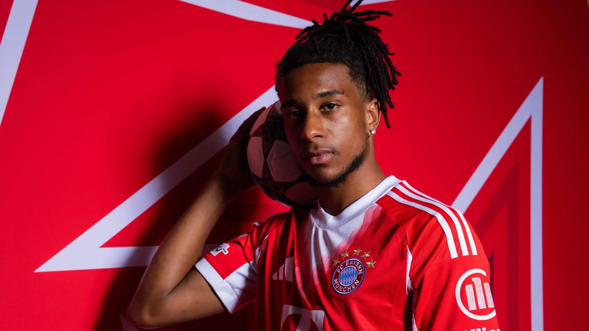 A player in a Bayern Munich jersey holding a football with a red background.