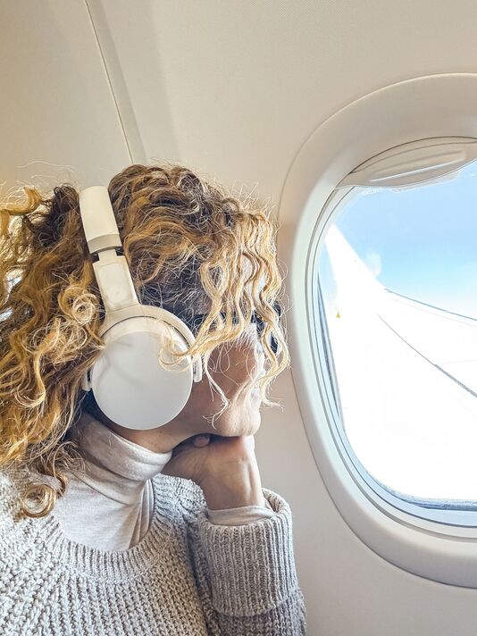 Woman with curly hair in noise-canceling headphones relaxes in an airplane window seat, gazing at blue sky and wing while enjoying music and peaceful 