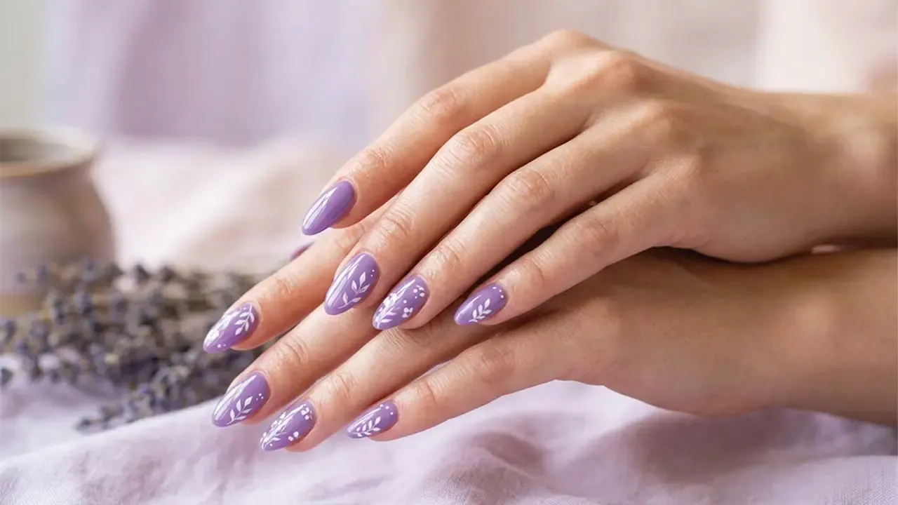 Woman displaying elegant purple manicured nails decorated with white leafy floral art placed over soft lavender fabric backdrop.