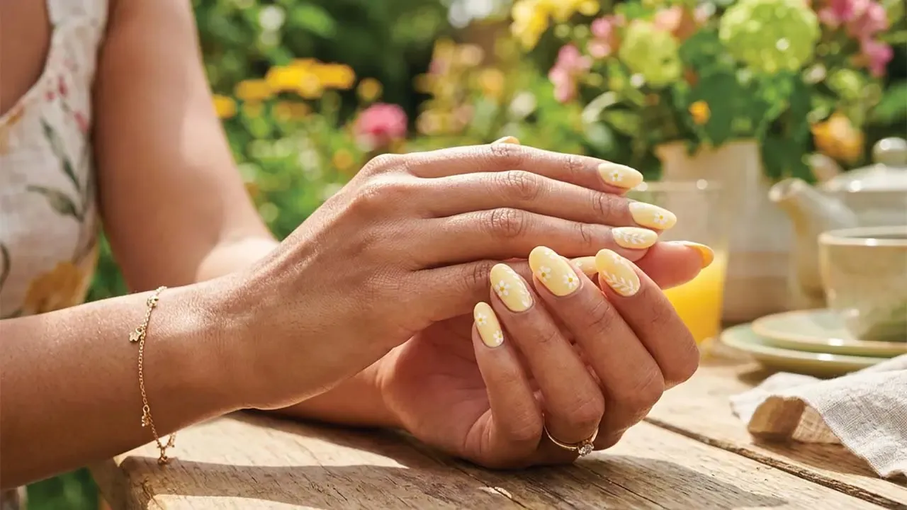 Woman displaying pastels yellow manicured nails featuring delicate white daisy floral art set in a sunny garden outdoor location.
