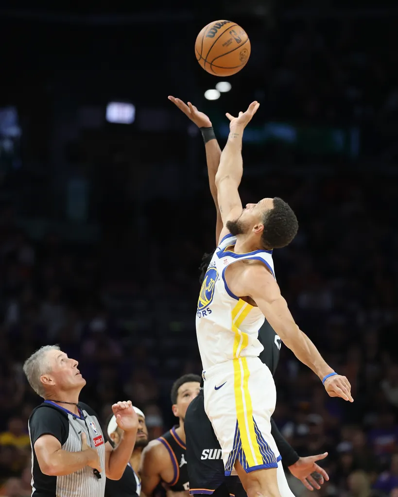 Basketball player in a white and gold uniform jumping for a jump ball against a dark background.