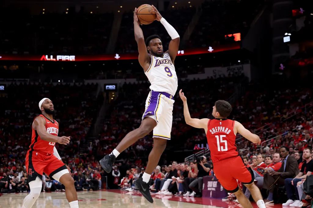 A Lakers player in a white and purple uniform jumps to shoot a basketball over a Rockets player in a red uniform.