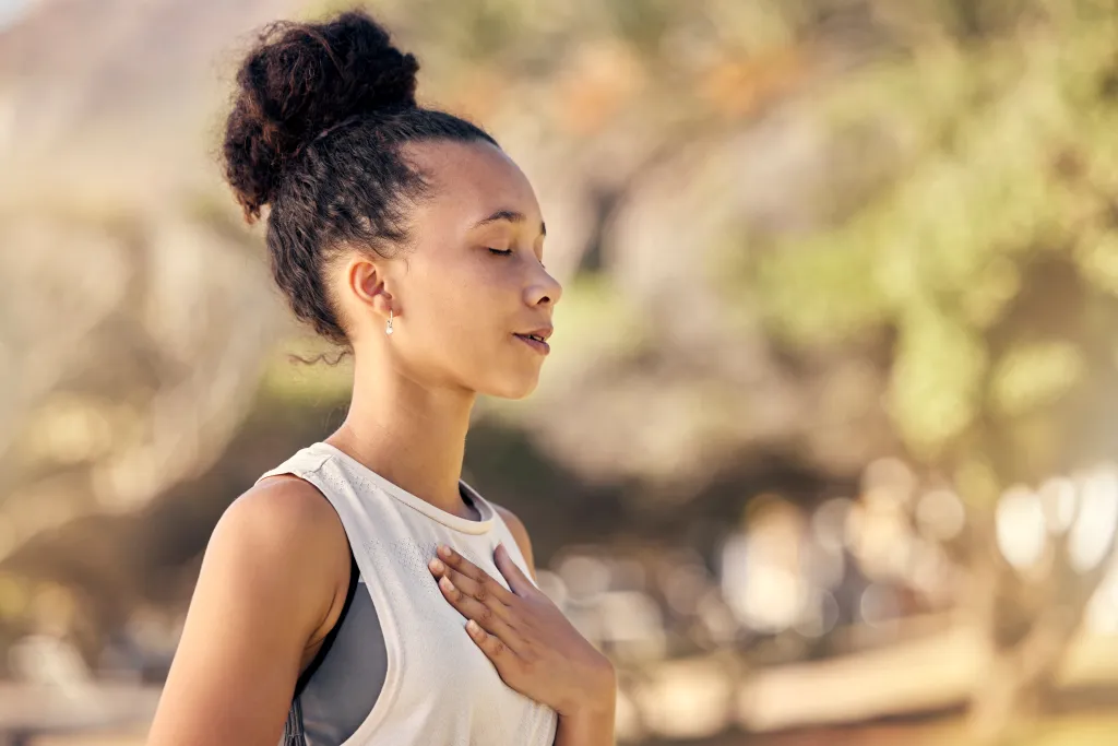 Young Black woman with eyes closed and hand on chest meditating outdoors.