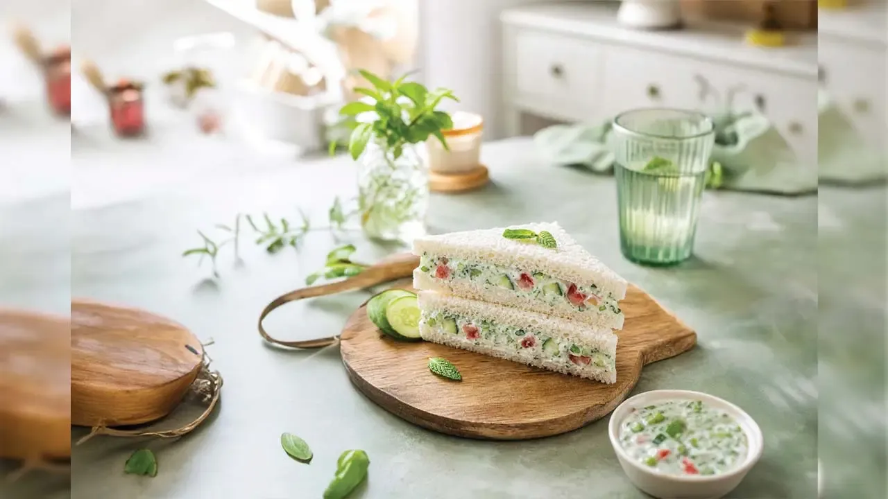Fresh creamy cucumber tomato sandwich slices served on wooden board with dip and water in bright setting.