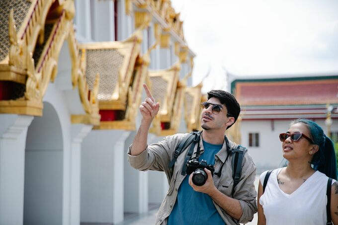 A visiting couple admires the architectural details of a traditional Thai temple, capturing the moment with a Canon camera. Photo by George Park via Pexels