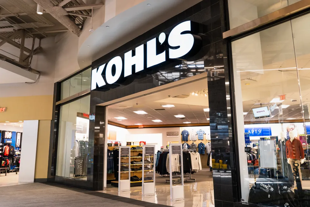 Exterior view of a Kohl's department store entrance with its illuminated logo and displays of clothing visible through the glass windows.
