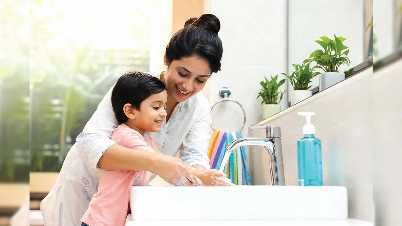 Smiling Indian mother helping young child wash hands with soap under running water in bright bathroom sink.