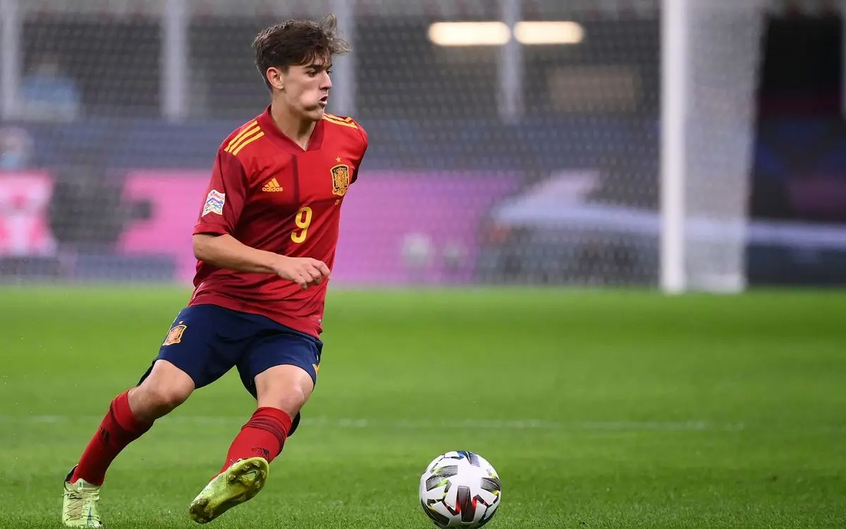Gavi in a red Spain national team kit dribbling a soccer ball on a green pitch.