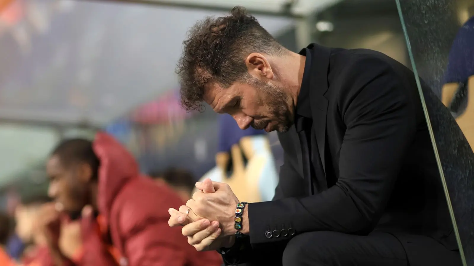 Diego Simeone sitting on the Atletico Madrid sideline wearing a black suit with a contemplative expression